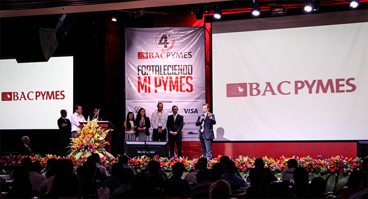 Evento corporativo de BAC PyMES con personas en el escenario frente a una audiencia y pantallas con el logotipo de la marca.