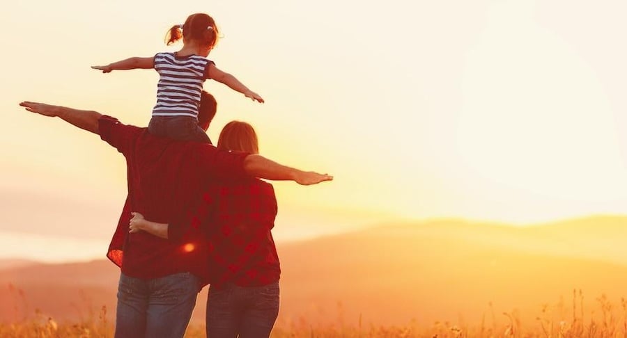 Vista posterior de una familia (padre, madre e hija) con los brazos extendidos frente a un atardecer, simbolizando salud y libertad.