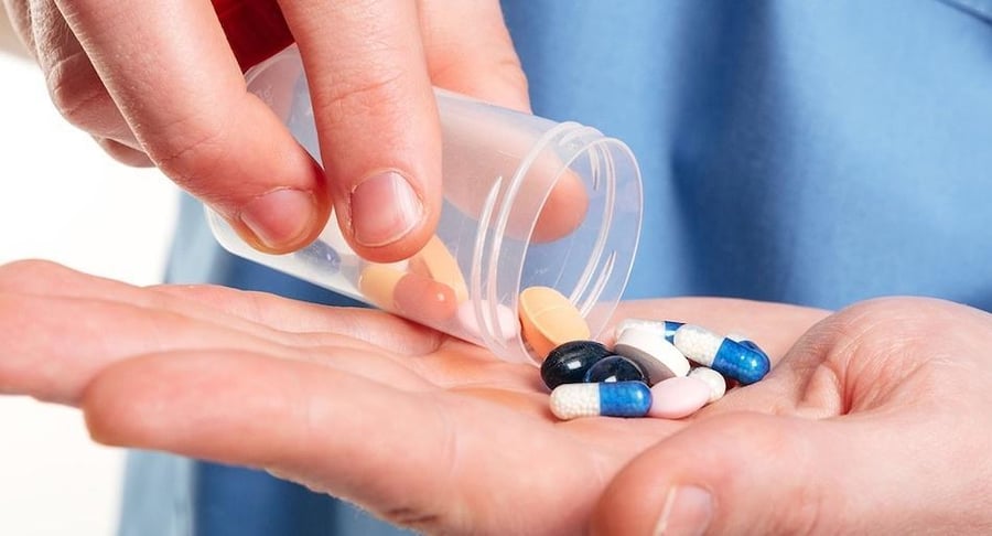 Primer plano de un profesional de la salud vertiendo cápsulas y pastillas de colores desde un frasco transparente hacia la palma de su mano.