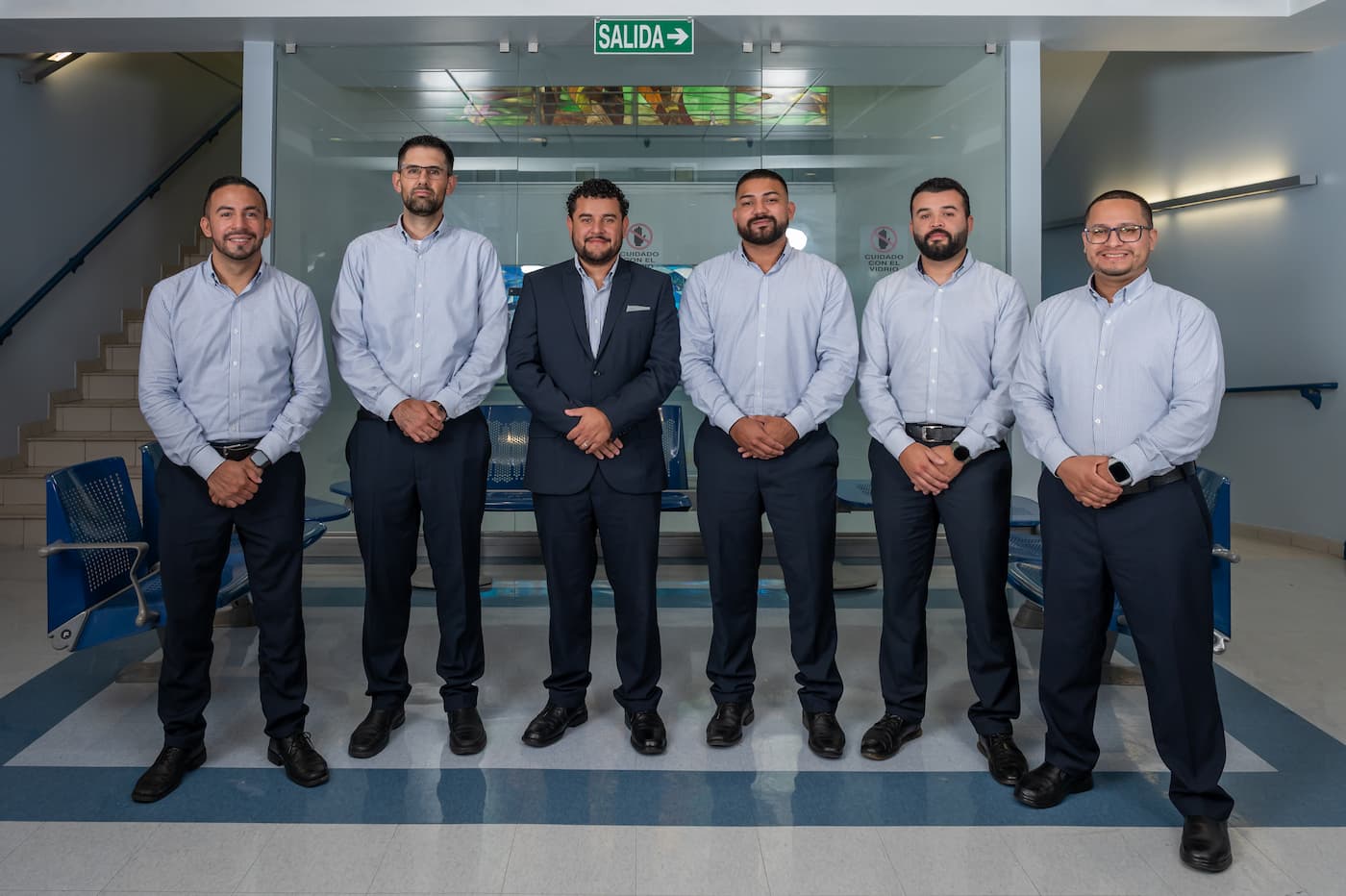 Foto de un grupo de seis hombres profesionales vistiendo camisa y traje formal, posando de pie en una sala de espera con sillas azuleadas y puertas de vidrio.