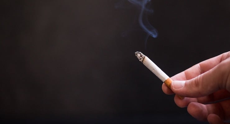 Mano sosteniendo un cigarrillo encendido con humo visible sobre fondo oscuro