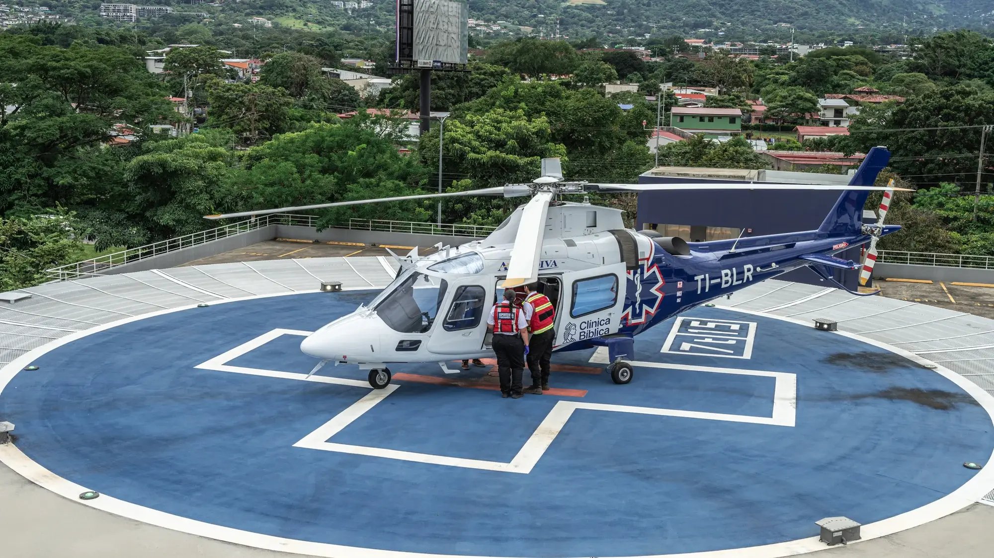 Blue and white helicopter of the Clínica Bíblica Hospital over a heliport surrounded by vegetation.