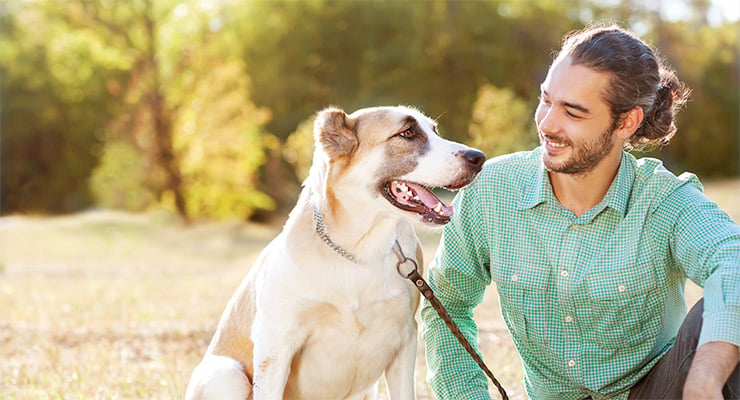 Hombre sentado al aire libre junto a su perro, ambos mirándose en un entorno natural.