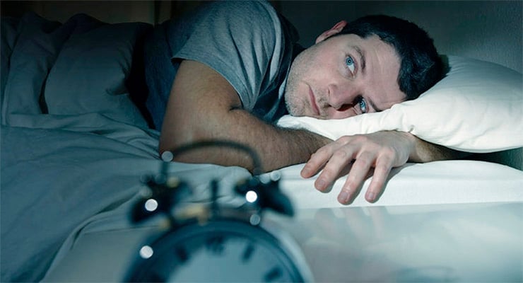 Hombre despierto en la cama durante la noche mirando un reloj despertador, con expresión de cansancio.
