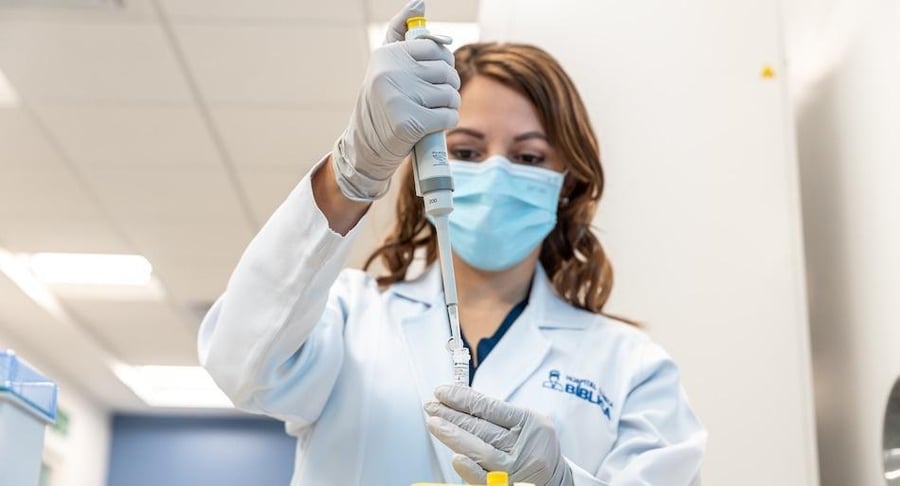 Profesional de laboratorio con bata y mascarilla utilizando una pipeta durante un análisis clínico.