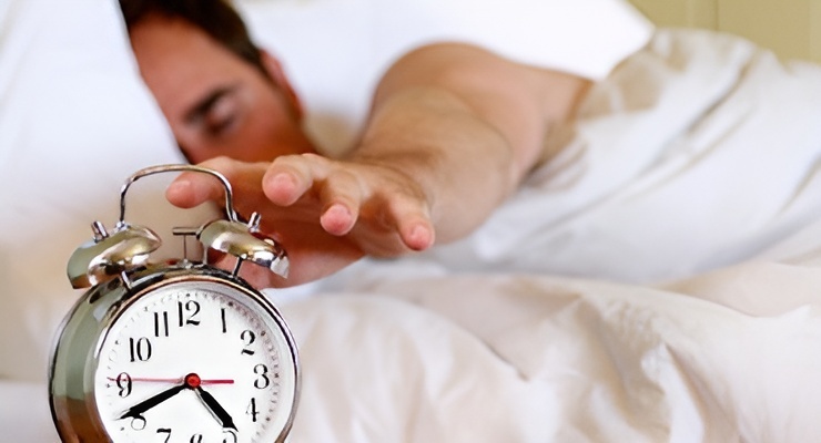 Persona en la cama estirando la mano para apagar un reloj despertador.