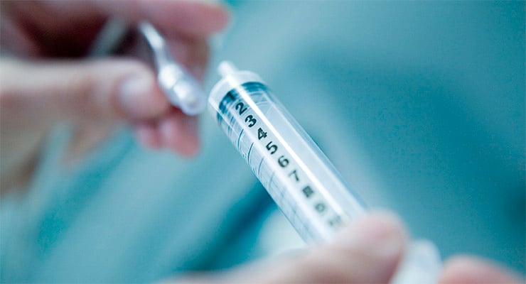 Jeringa preparada para vacunación sostenida por manos con un frasco de medicamento al fondo.