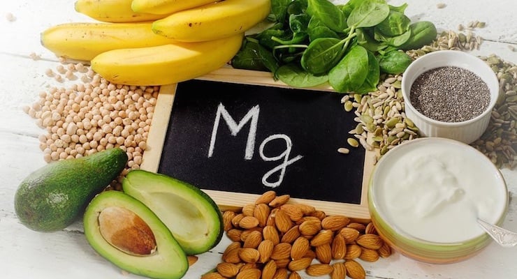 Alimentos ricos en magnesio como aguacate, banano, almendras, semillas y yogur alrededor de pizarra con símbolo Mg