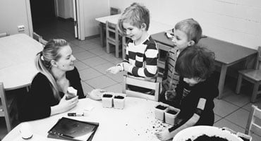 Docente conversando con varios niños alrededor de una mesa con recipientes y material educativo en un aula