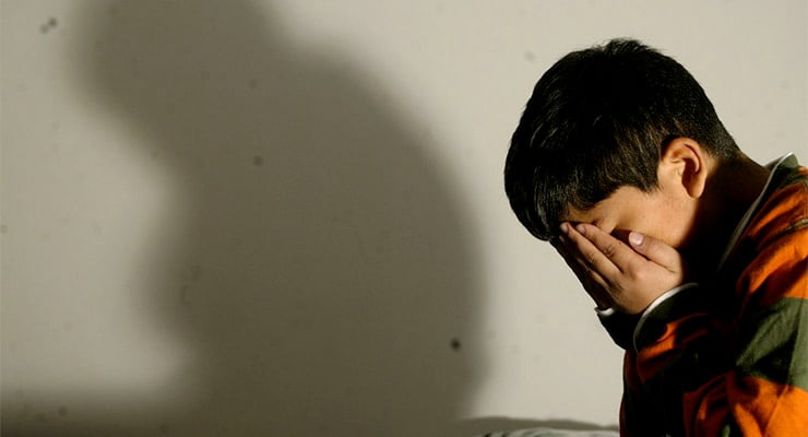 Niño sentado con el rostro cubierto por las manos y una sombra proyectada en la pared