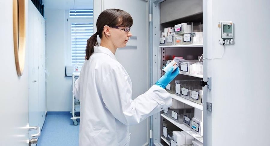 Farmacéutica con bata blanca y guantes azules revisando el inventario dentro de un refrigerador médico con control de temperatura digital.