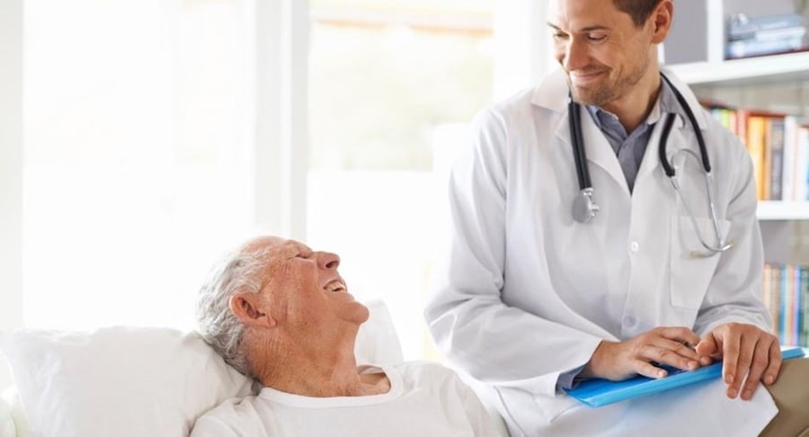 Un médico sonriente con bata blanca y estetoscopio conversa con un paciente adulto mayor que descansa en una cama, representando el acompañamiento en salud.