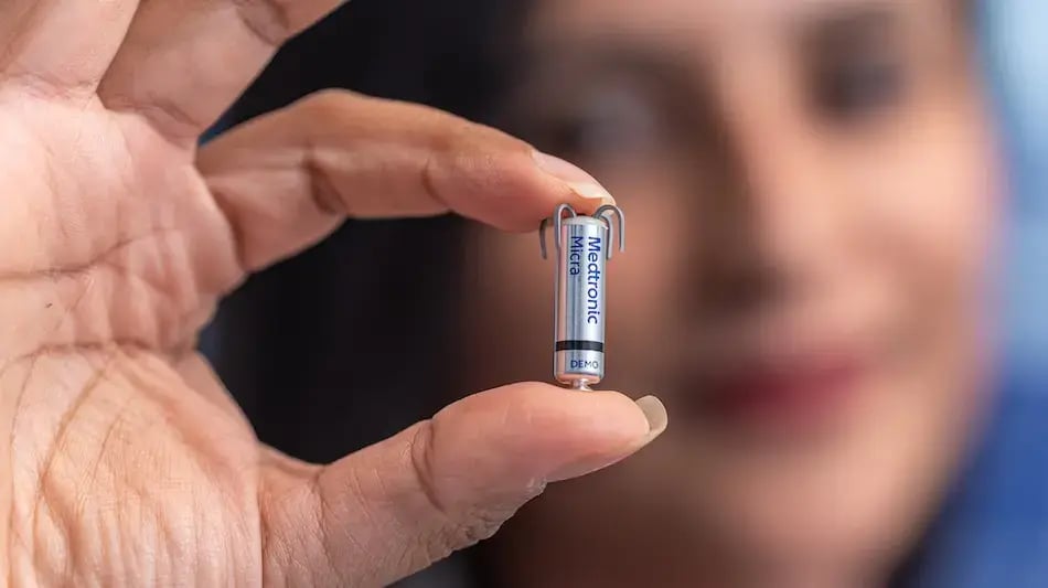 Mano sosteniendo un pequeño dispositivo de marcapasos frente a un rostro desenfocado.