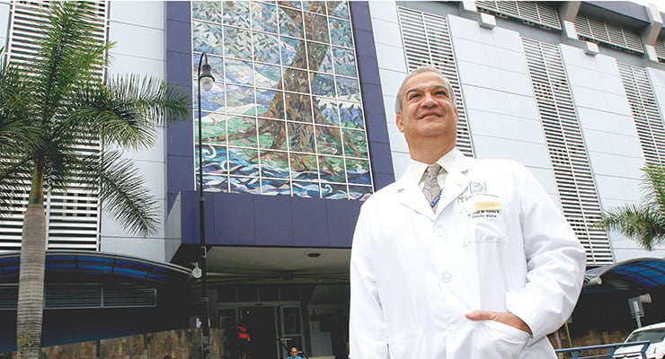 Médico con bata blanca de pie frente al edificio de la Clínica Bíblica