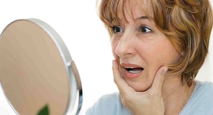 Mujer adulta observándose el rostro frente a un espejo con expresión de sorpresa.