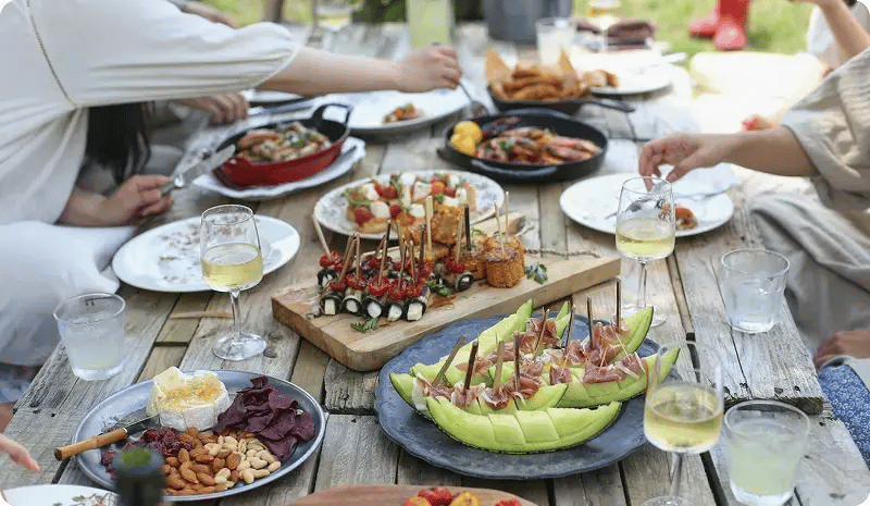 Mesa de madera al aire libre con diversos platillos servidos y varias personas alrededor alcanzando la comida.