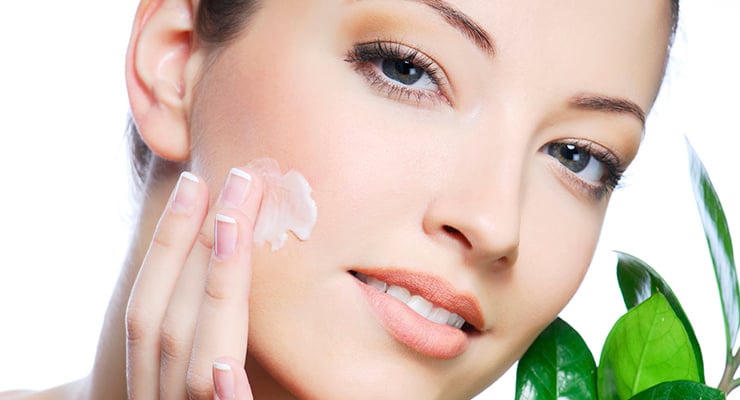 Mujer aplicando crema facial en la mejilla con hojas verdes al fondo