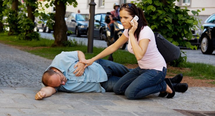 Mujer arrodillada auxiliando a un hombre que yace en el suelo mientras habla por teléfono en la vía pública.