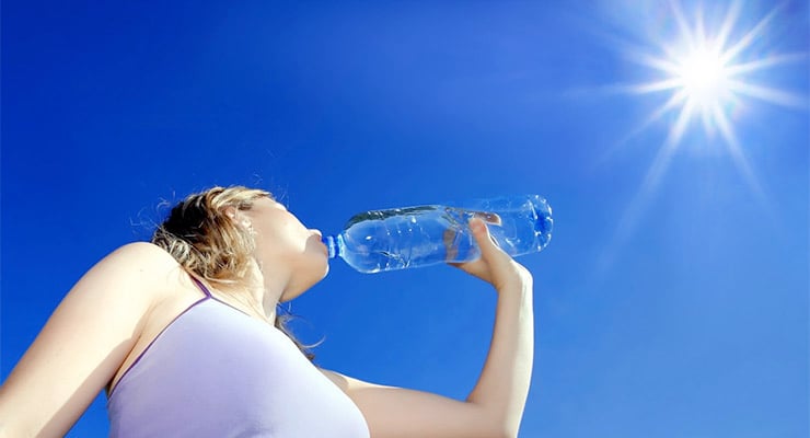 Mujer bebiendo agua de una botella bajo el sol intenso en un cielo despejado.