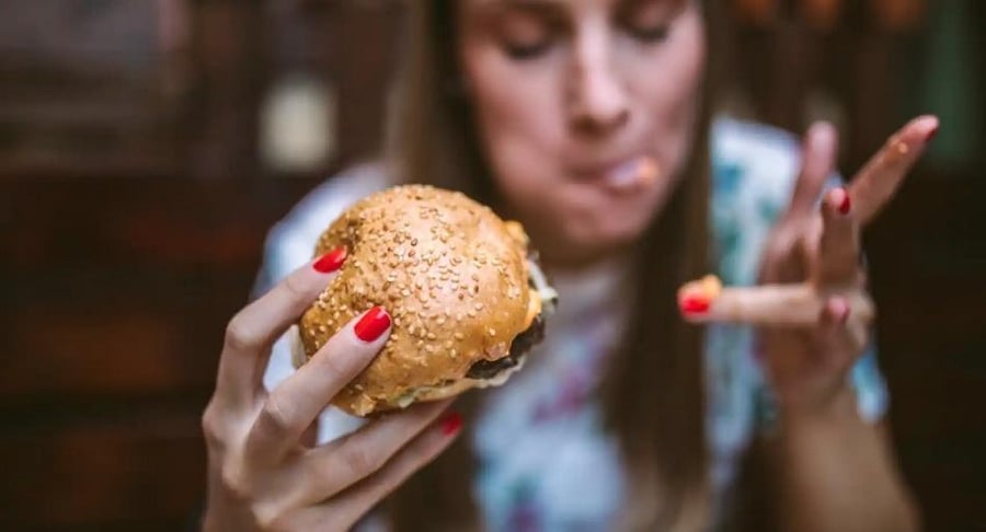Primer plano de una mujer sosteniendo una hamburguesa y comiendo con ansiedad, representando el concepto de hambre emocional.