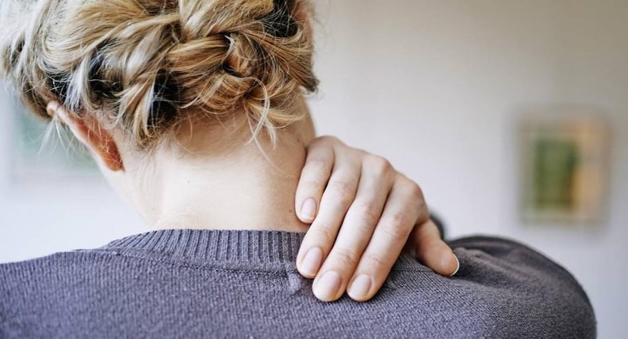 Vista posterior de una mujer con el cabello recogido sujetando su cuello con la mano, expresando dolor físico derivado de tensión emocional.