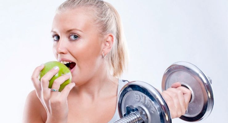 Mujer sosteniendo una mancuerna mientras come una manzana verde en un entorno de ejercicio.