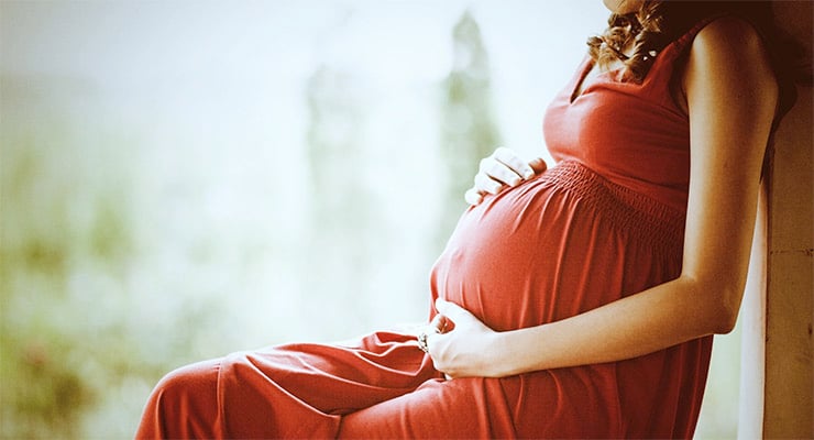 Mujer embarazada sentada, con vestido rojo, apoyando las manos sobre el abdomen.