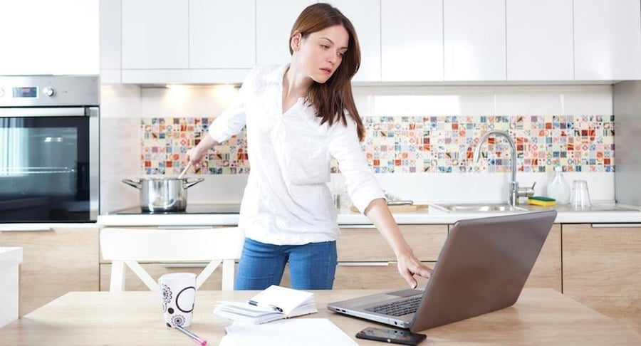 Mujer joven en una cocina moderna trabajando con su computadora portátil mientras supervisa una olla en la cocina, representando el reto del balance vida-trabajo.