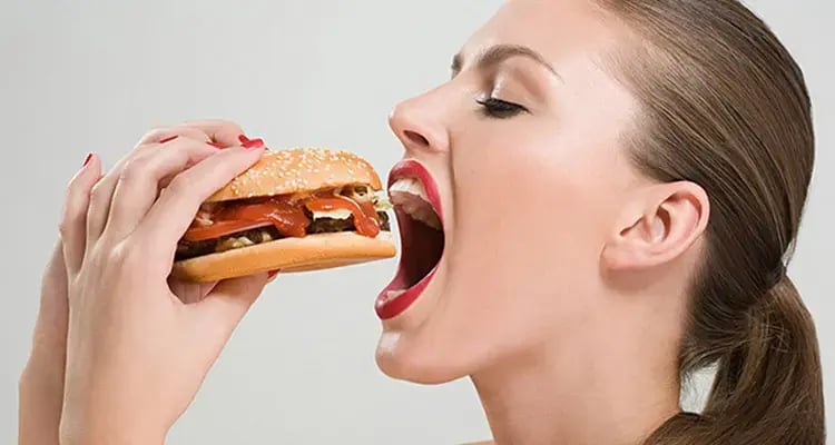 Mujer sosteniendo y acercando a su boca una hamburguesa con salsa