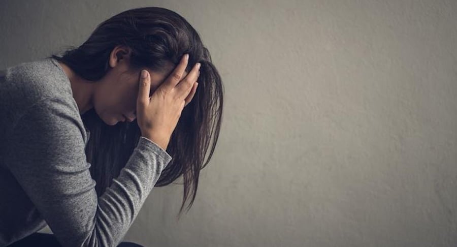 Perfil de una mujer joven con el rostro cubierto por sus manos en un gesto de agotamiento o estrés, sobre un fondo gris neutro.