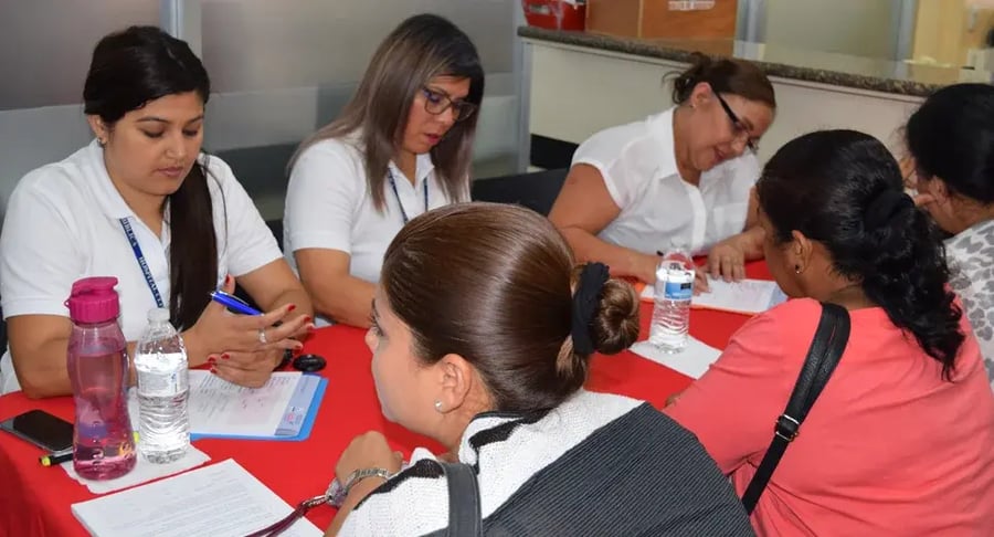 Mujeres sentadas en una mesa mientras completan formularios y conversan con personal administrativo.