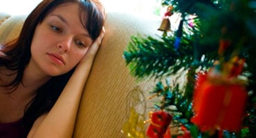 Mujer joven con expresión de melancolía y tristeza sentada en un sillón junto a un árbol de Navidad decorado.