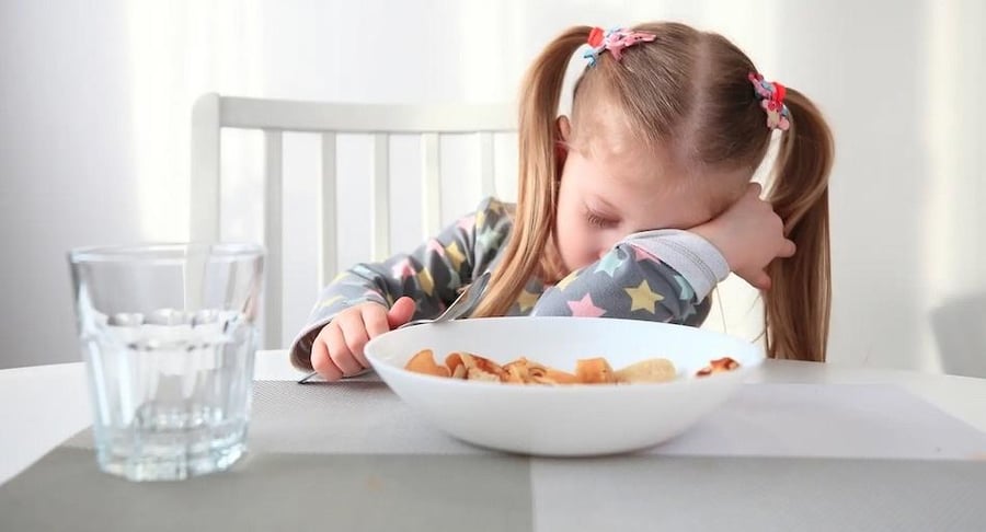 Niña pequeña con coletas apoyando su cabeza sobre el brazo en la mesa, mostrando signos de cansancio y debilidad por anemia.