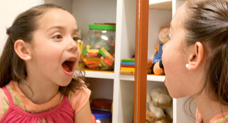 Niña frente a un espejo realizando ejercicios de pronunciación en un espacio con juguetes educativos.