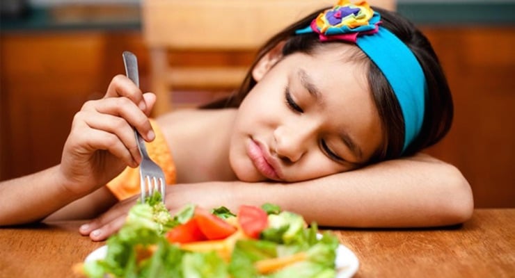 Niña sentada a la mesa con plato de ensalada mientras muestra falta de apetito