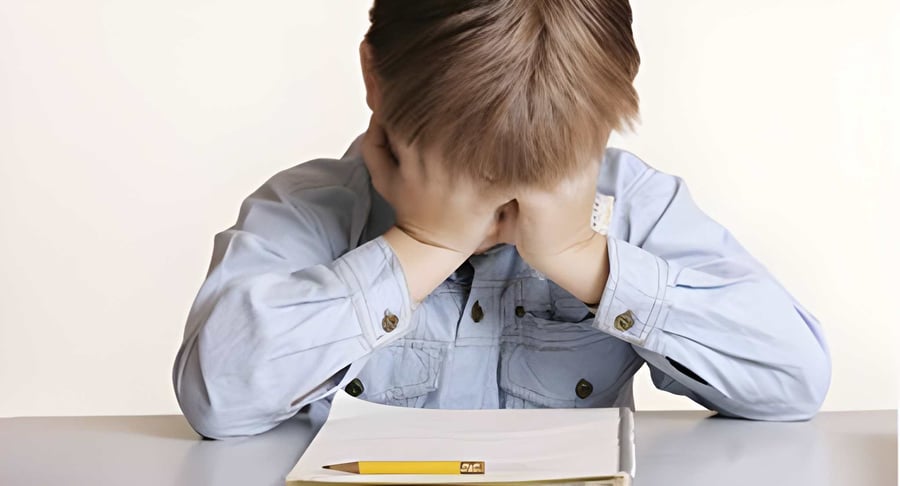 Niño sentado frente a una mesa con la cabeza cubierta por las manos y un cuaderno al frente