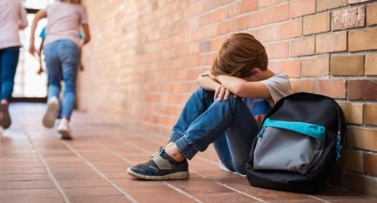 Niño sentado en el suelo con mochila mientras otros niños se alejan en un pasillo escolar