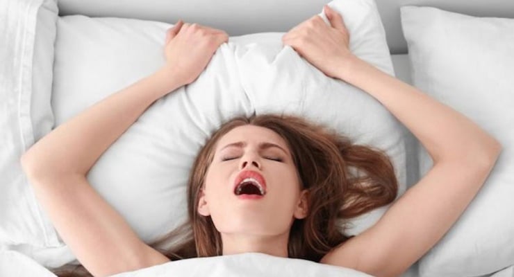 Vista superior de una mujer joven en una cama con sábanas blancas, con los ojos cerrados y expresión de placer, representando el orgasmo femenino.