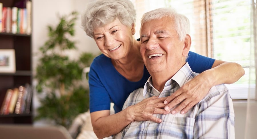 Pareja de adultos mayores sonríe y se abraza en el hogar, representando bienestar emocional y un estilo de vida saludable en la adultez mayor.
