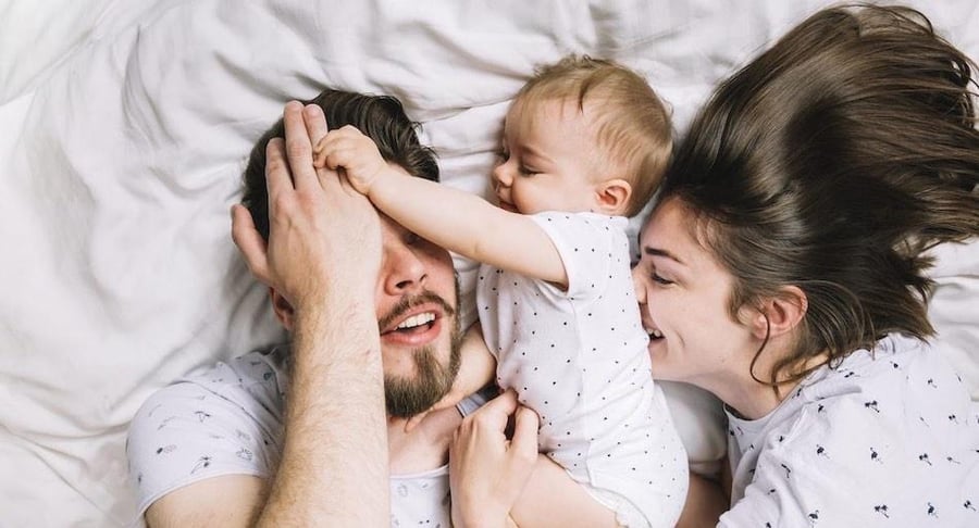 Pareja joven con un niño pequeño jugando alegremente en una cama con sábanas blancas, representando la planificación familiar y felicidad.