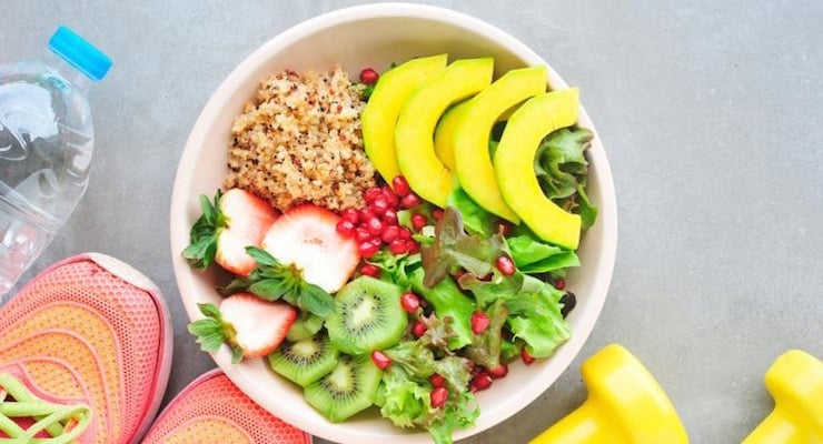 Plato con ensalada saludable, quinoa, aguacate, frutas y vegetales junto a pesas y botella de agua