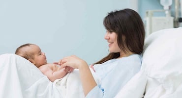 Madre en cama hospitalaria observando a su bebé recién nacido con expresión tranquila