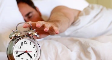 Persona acostada en una cama extendiendo la mano hacia un despertador