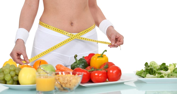 Persona midiendo su cintura con cinta métrica frente a alimentos saludables como frutas y vegetales