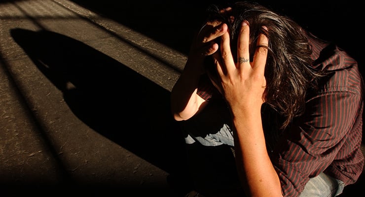 Persona sentada en el suelo con la cabeza entre las manos, proyectando una sombra alargada en un espacio con luz tenue.