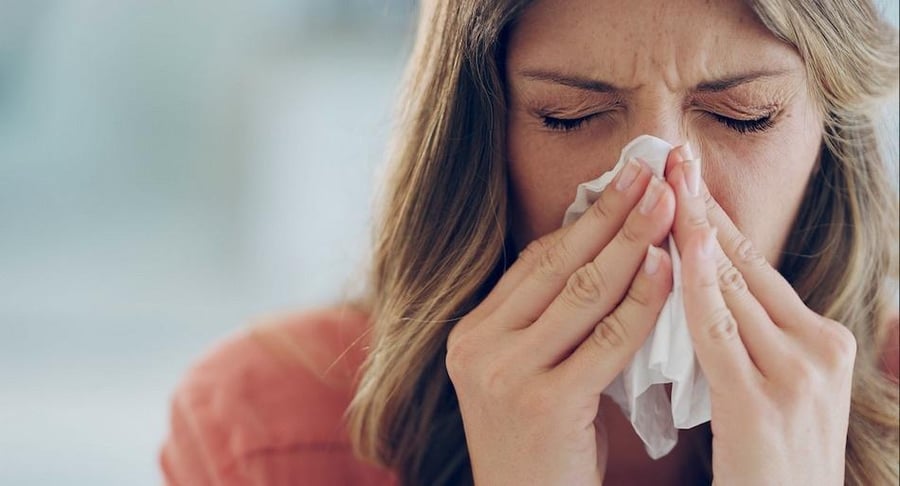 Primer plano de una mujer con los ojos cerrados utilizando un pañuelo de papel para sonarse la nariz, representando síntomas de rinitis o alergia.