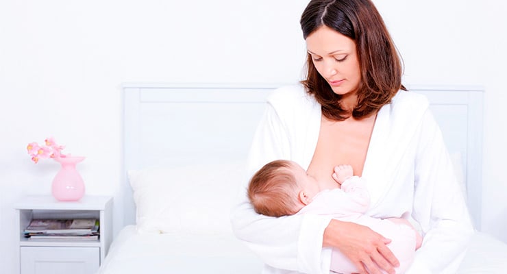 Madre amamantando a su bebé sentada en una cama en un entorno claro