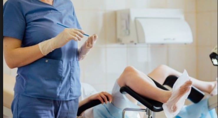 Una profesional de la salud con uniforme azul y guantes blancos prepara el equipo médico en un consultorio ginecológico para un examen de Papanicolau.