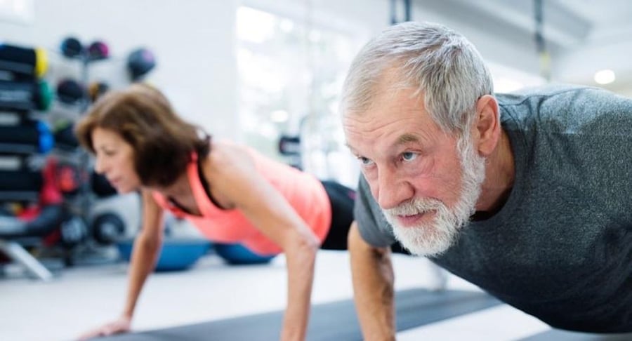 Un hombre y una mujer adultos mayores realizando ejercicios de plancha sobre colchonetas en un gimnasio para fortalecer sus músculos.