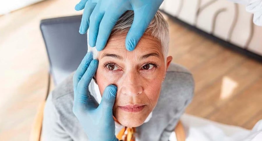 Un oftalmólogo con guantes azules realiza un examen visual a una paciente adulta mayor, revisando cuidadosamente el área de sus ojos.